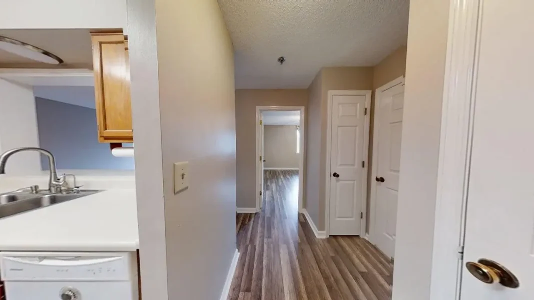 Hallway of a home with beige walls, wood-look flooring, and white doors, viewed from a kitchen area toward a room at the end