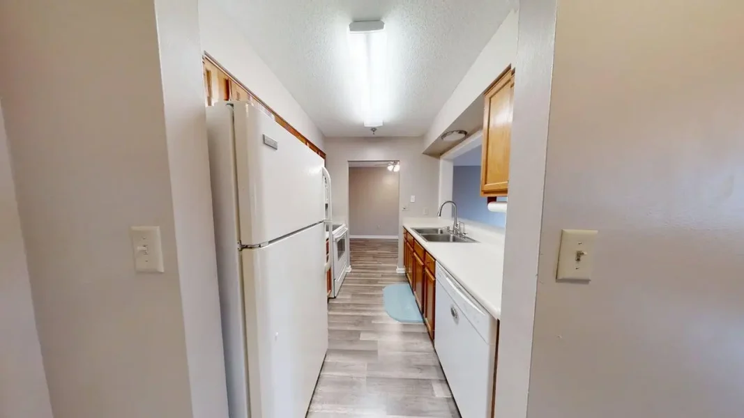 Narrow galley kitchen with a white refrigerator on the left, white countertops, wooden cabinets, a double sink, and a light gray floor.