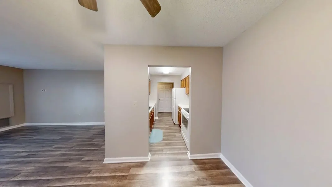 Open living area with wood floors and beige walls, opening into a small kitchen with white appliances and wooden cabinets.