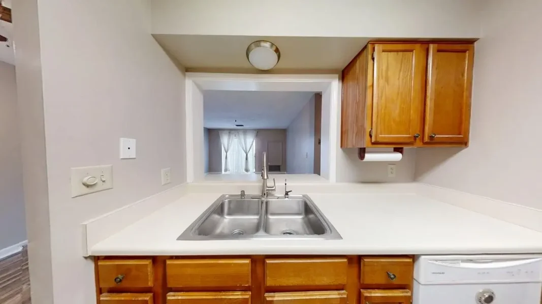 Kitchen with a double stainless steel sink, white counters, and oak cabinets; a pass-through opening reveals the living room beyond.