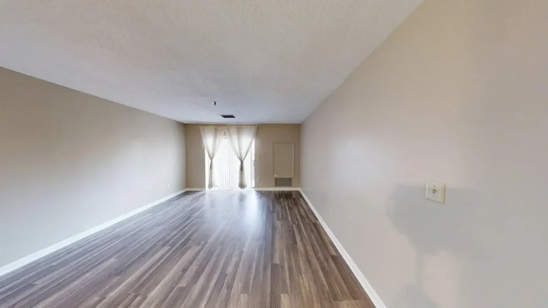 Empty living room with light gray wood flooring, beige walls, and sheer curtains at the back door. short and bright space for entertaining or relaxing.