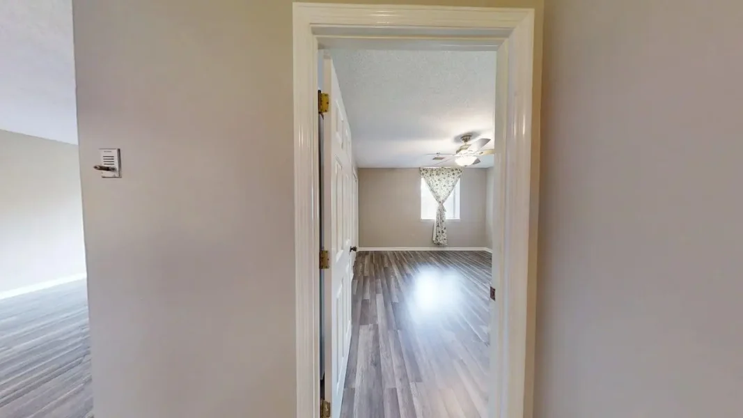 Empty room with gray wood-look floors, a window with floral curtain, and a ceiling fan, seen from an open doorway.