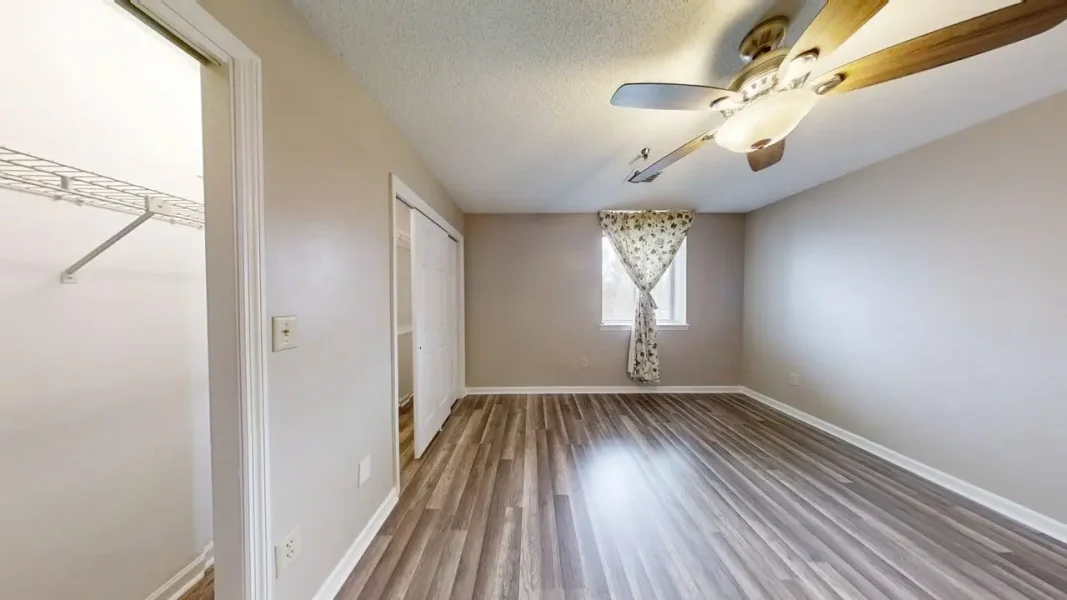 Empty bedroom with wood-look laminate flooring, beige walls, and a ceiling fan; a window with a floral curtain lets in light.