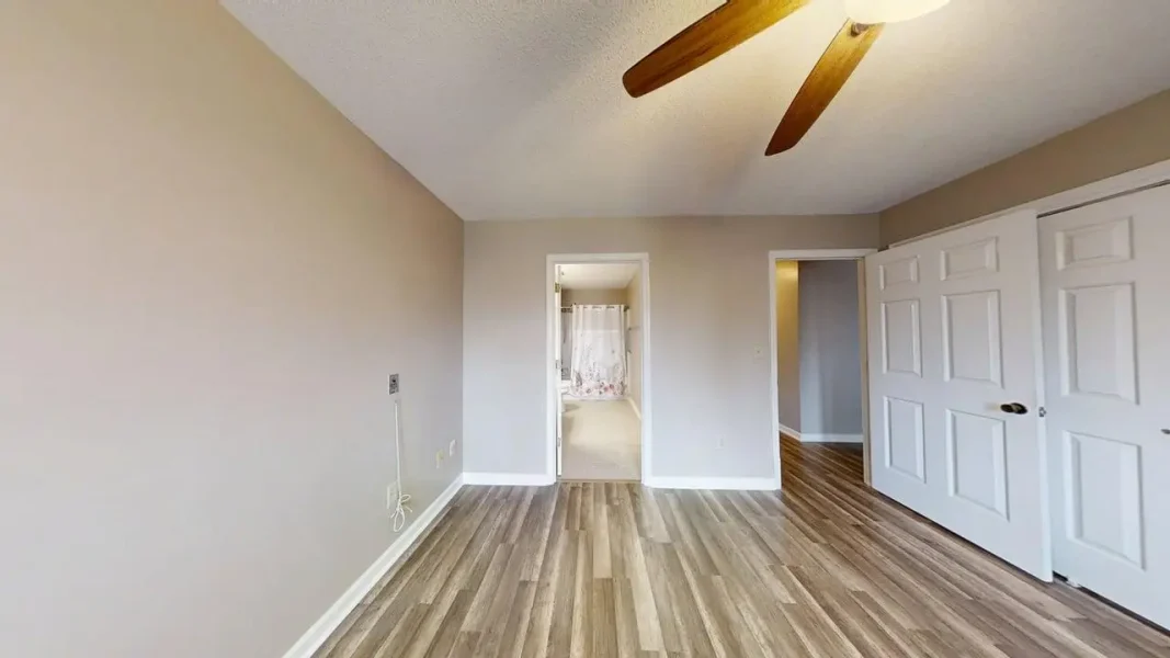Empty bedroom with wood-look flooring, beige walls, a ceiling fan, and white double closet doors on the right; open doorway reveals a bathroom ahead with a shower curtain visible.