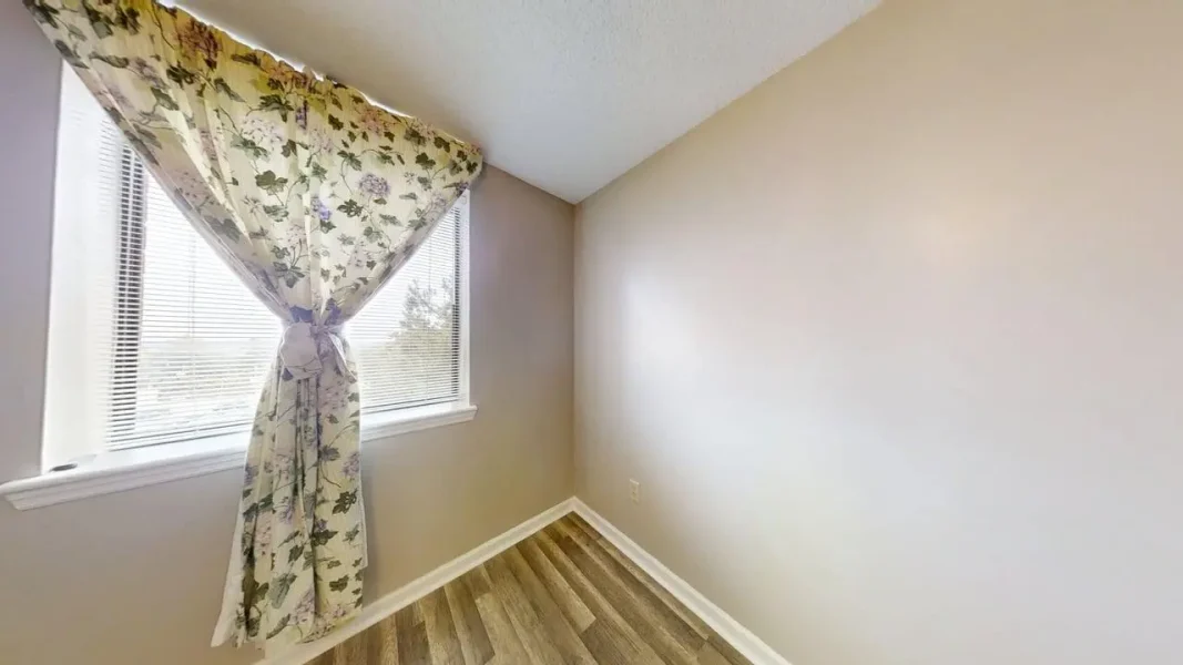 Small empty corner room with beige walls, wood-looking flooring, and a floral curtain tied by the window.