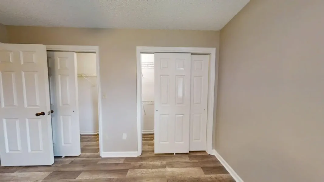 Empty beige room with light wood flooring, two white doors—one open to a closet with wire shelves—and a sliding closet door in the center.