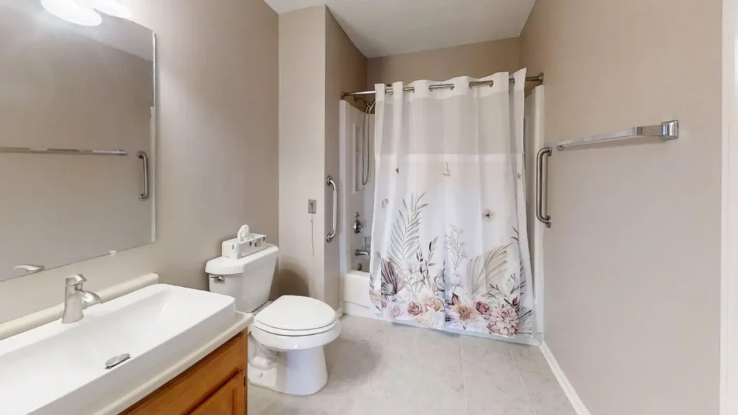 Bathroom with a white sink and wooden vanity, large wall mirror, and a toilet next to a bathtub with a plant-themed shower curtain; includes accessibility grab bars on the walls for support.
