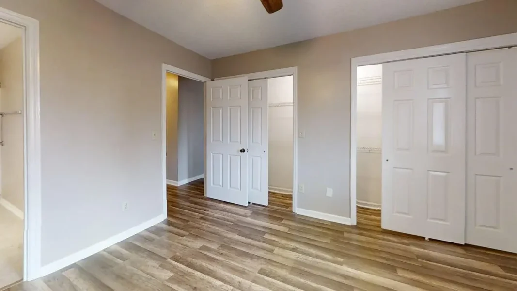 Empty bedroom with beige walls, white baseboards, and wood-look laminate flooring; two white closet doors on the right and an open doorway ahead.”​,