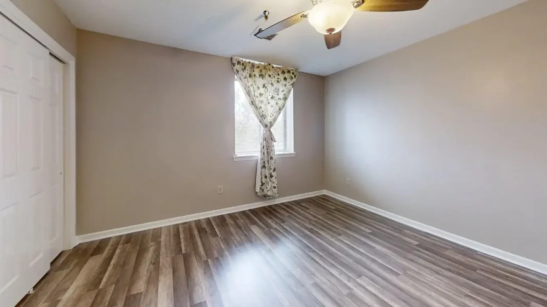 Empty bedroom with beige walls, white baseboards, window with floral tie-back curtain, ceiling fan, and wood-look flooring. A tidy, unfurnished room with a closet door on the left.