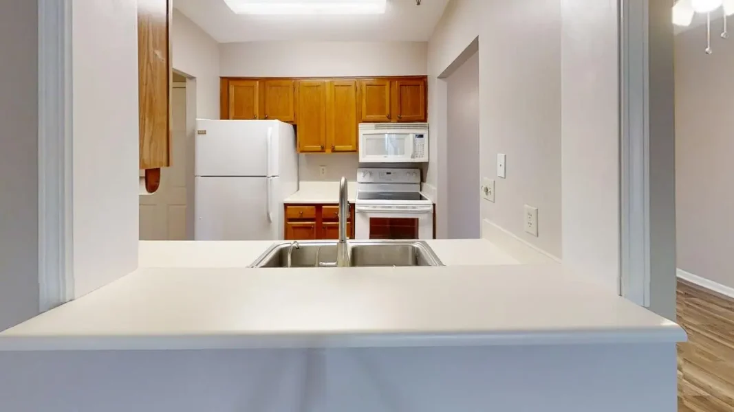 Double-sink kitchen with white countertops, wooden cabinets, and white fridge, microwave, and stove visible in the background.