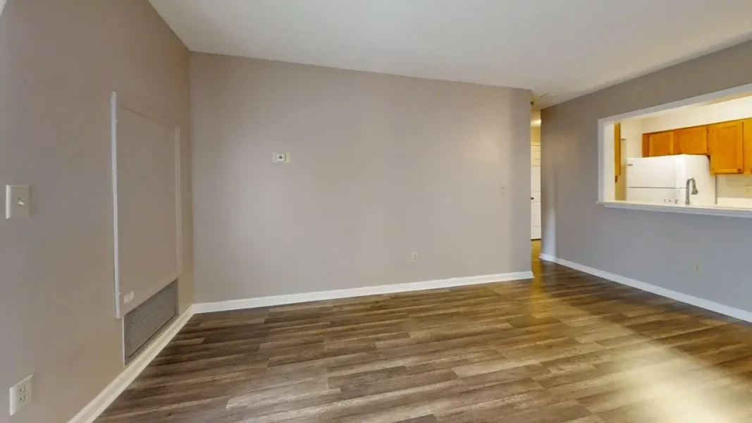 Empty living room with light gray walls, wood laminate flooring, and a kitchen pass-through on the right. A vent and thermostat visible on the left wall.