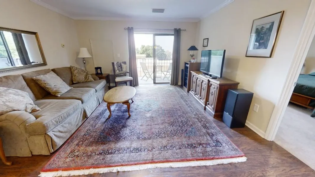 Living room with a beige sofa, throw pillows, and a wooden coffee table on a large patterned area rug by a sliding glass door to a balcony.