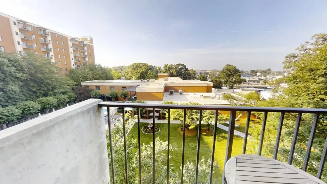 Balcony railing frames a grassy courtyard with trees and low yellow-brick buildings in the distance on a sunny day.