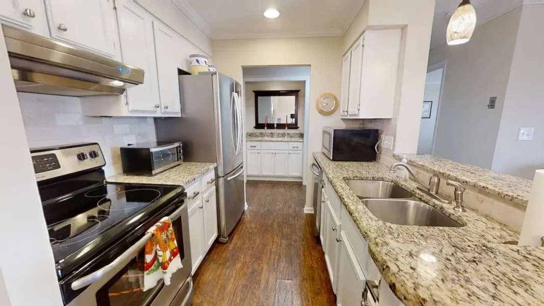 Bright galley kitchen with white cabinets, granite counters, and stainless steel fridge, stove, and double sink island in the center.