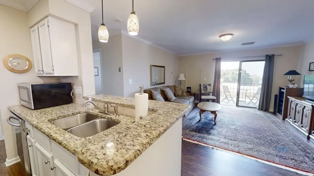 Open-concept living room with a granite kitchen island, double sink, microwave, and pendant lights, leading to a seating area and balcony.