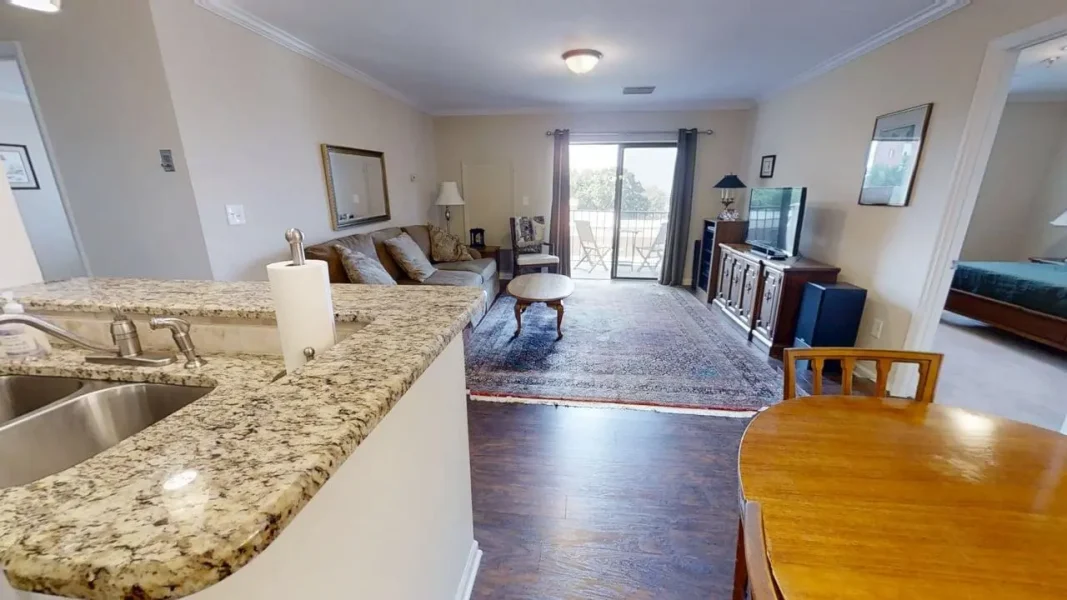 Open-concept living area with a granite kitchen counter in the foreground, sink, and paper towel roll, leading to a beige sofa and coffee table on a rug. A sliding door to a balcony is in back.