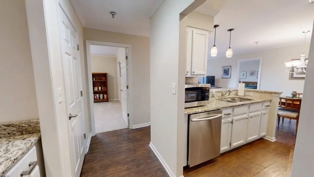Open-concept kitchen with granite countertops, white cabinets, stainless steel dishwasher and microwave, and an island; hallway leads to a living area.