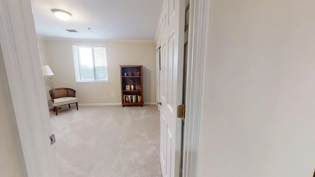 Light-filled living room seen from doorway: beige walls, window with blinds, wooden bookshelf, upholstered chair, and beige carpet.