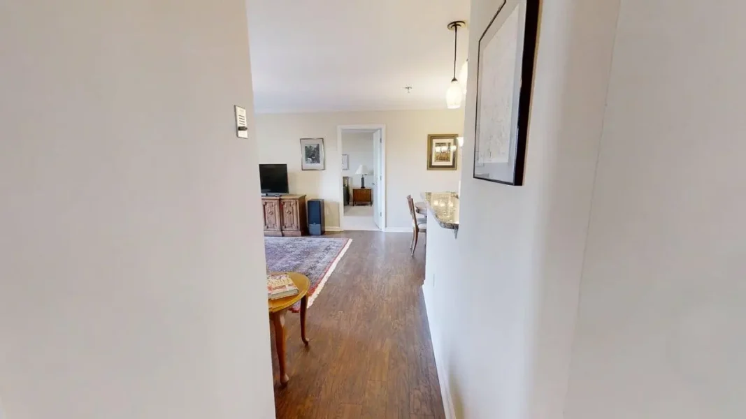 View down a hallway into a living room with a TV on a wooden cabinet, a blue rug, and a doorway to another room; a dining area is visible on the right.