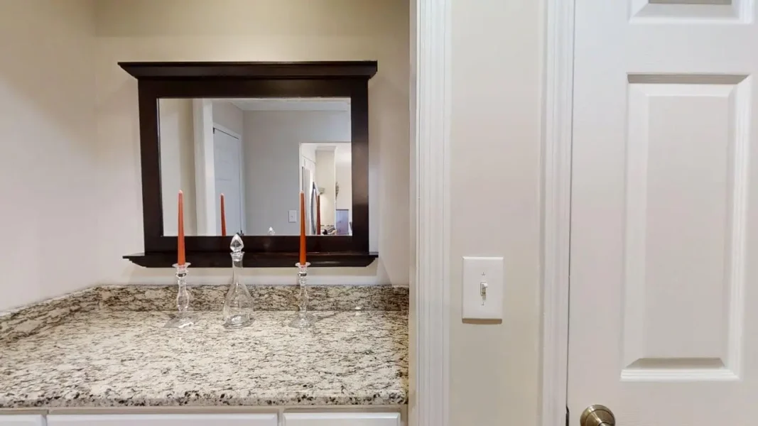 Dark-framed mirror above a granite vanity with two crystal candle holders and orange candles, reflection of a hallway in the mirror.