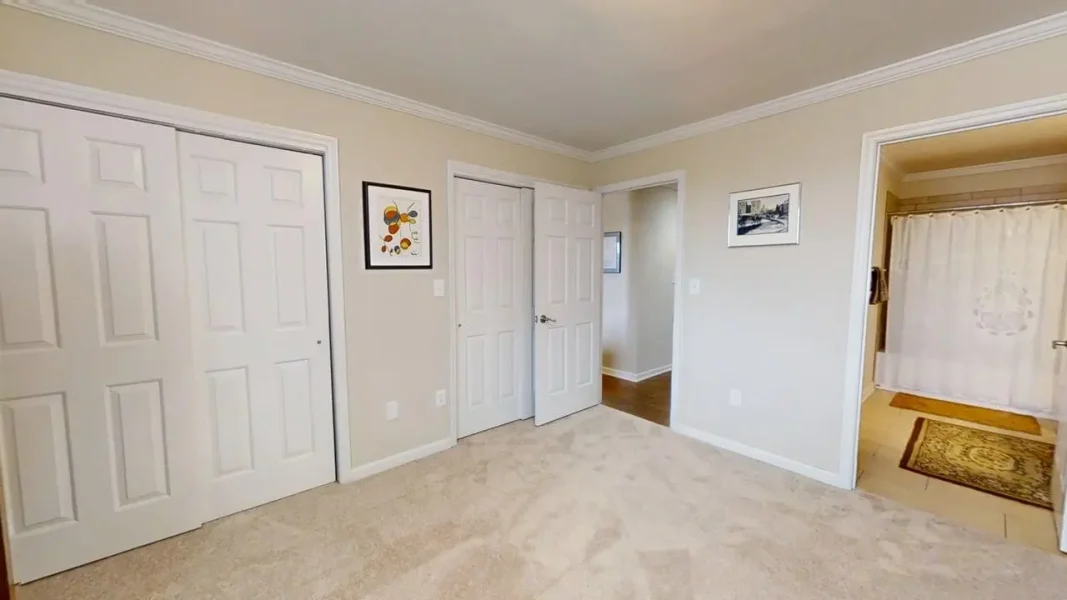 Bedroom with beige carpet and two large white closet doors on the left, doorway to hall in center, and bathroom with white shower curtain on the right.