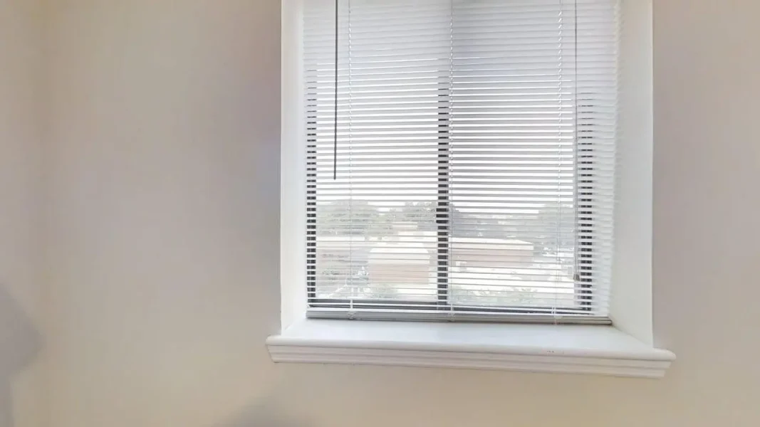 Window with white horizontal blinds partially open, letting light into the room behind a white sill.