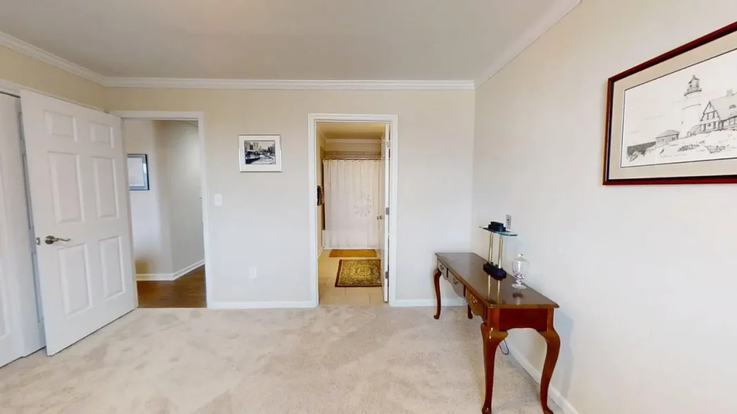 A bedroom with a beige carpet, white doors, and a wooden console table against the right wall, framed picture above.