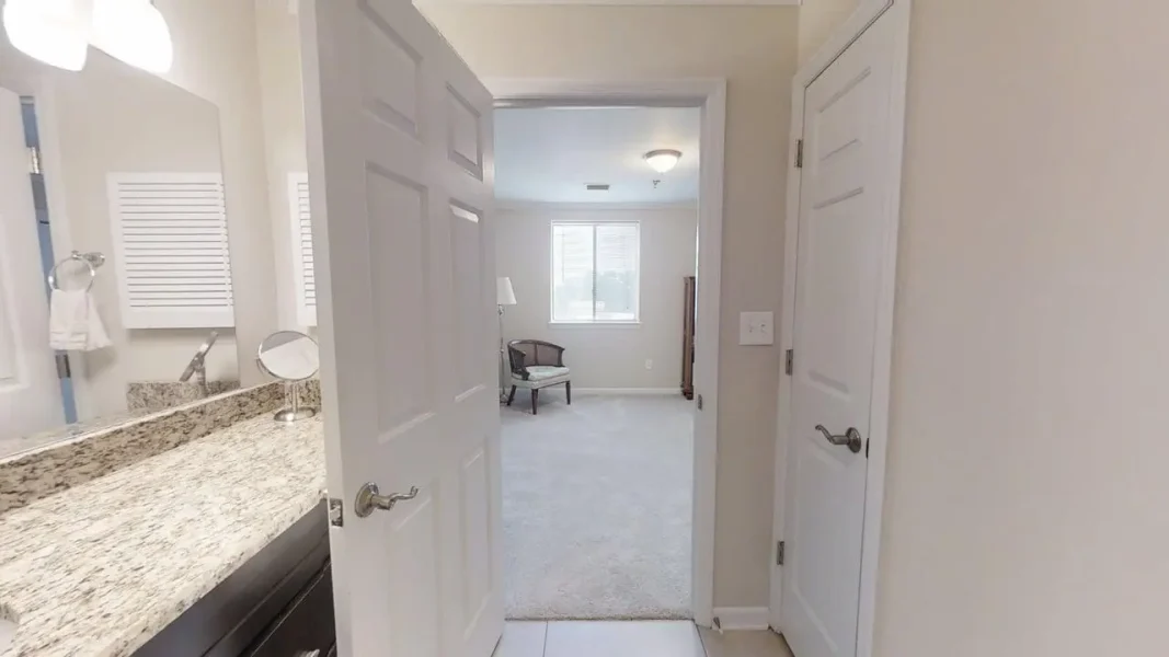 View from a bathroom into a carpeted living area; granite countertop and sink on the left, open white doors frame a sunlit room with a chair by the window in the distance.