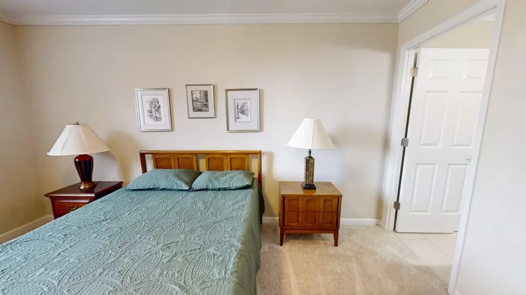 Bedroom with a wooden bed frame, teal quilt, and two matching nightstands with lamps on each side. Three framed pictures hang above the headboard on beige walls.