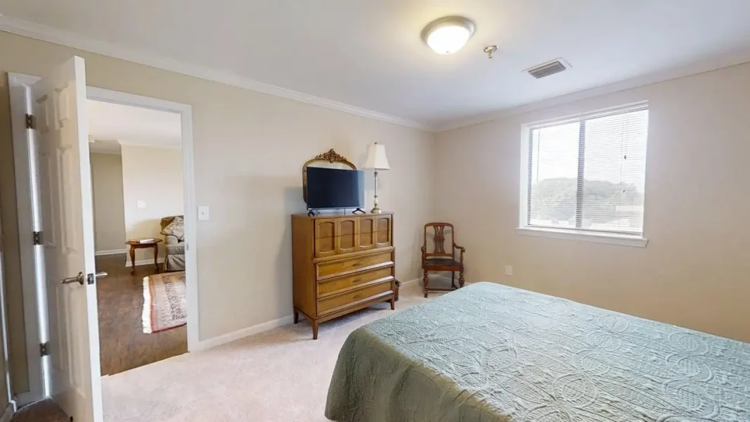 Bedroom with a bed covered in a light blue quilt, wooden dresser with TV and lamp, and a vintage chair by a window.