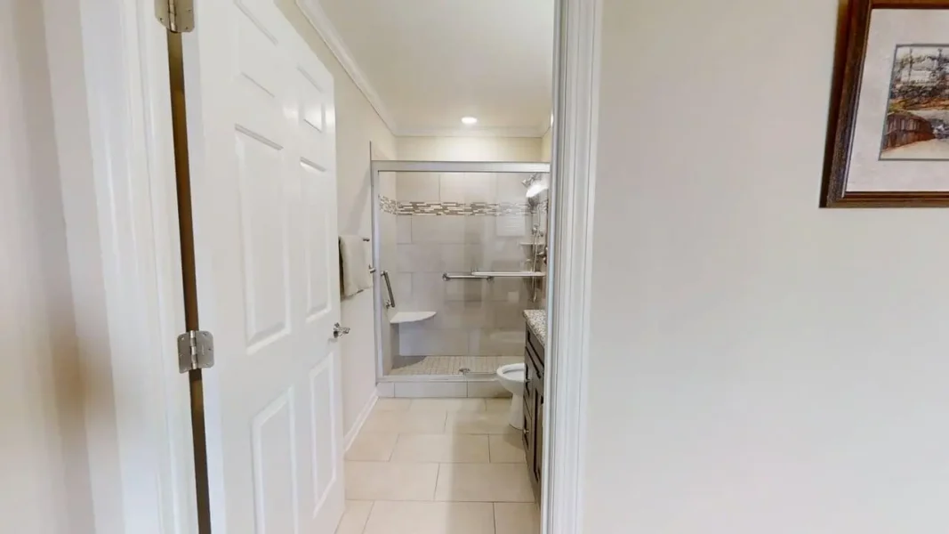 Bright bathroom seen through an open doorway: glass-enclosed shower with grab bars, tiled walls and floor, towel on rack, toilet and dark vanity with granite countertop to the right.