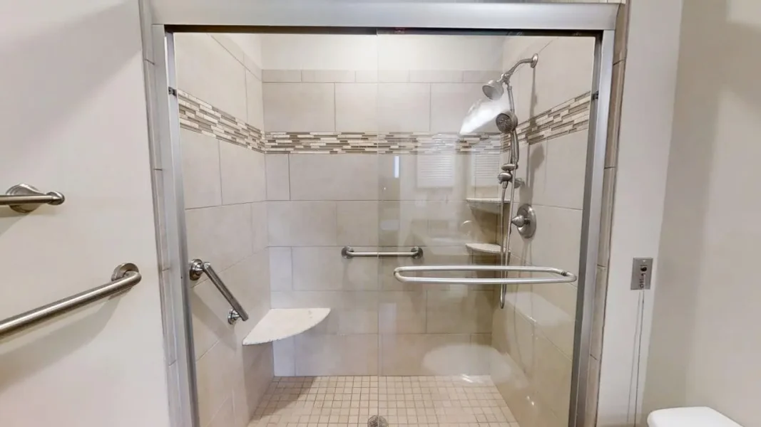Accessible glass-enclosed shower stall with built-in seat, horizontal grab bars, and a handheld showerhead.