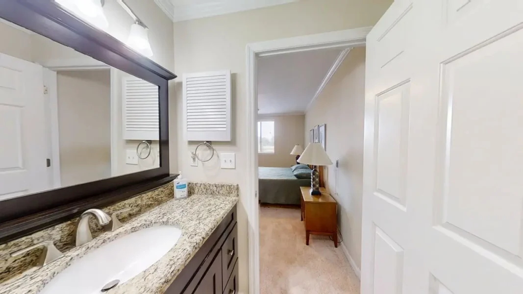 Bathroom vanity with granite countertop and double sink, view toward an adjoining bedroom with a bed and nightstand.