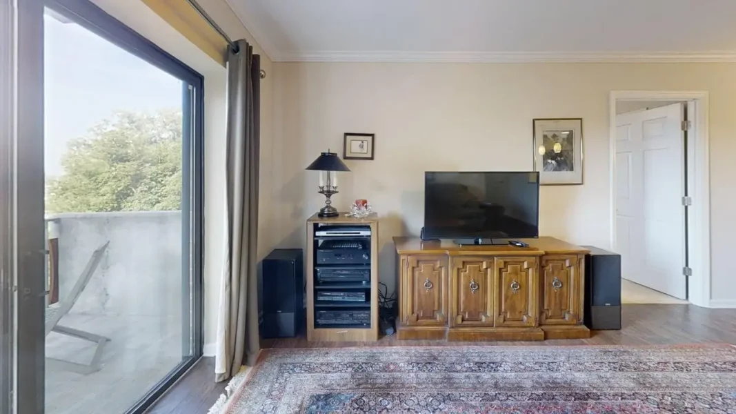 Living room with a flat-screen TV on a carved wooden console, stereo components, and a table lamp near a sliding glass door to a balcony.