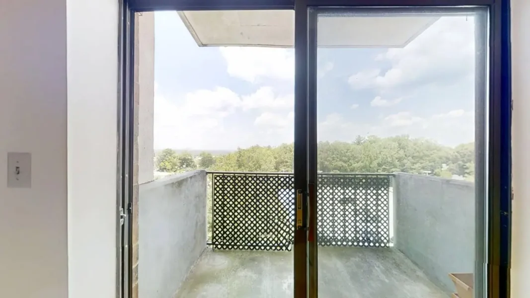 Sliding glass door opens to a small balcony with a perforated metal railing, overlooking trees and a blue sky.