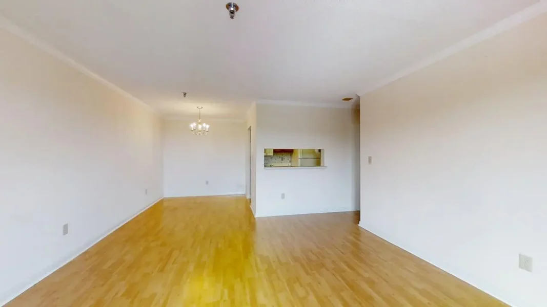 Empty living room with light wood floors, white walls, and a kitchen pass-through window; chandelier visible in the distance.