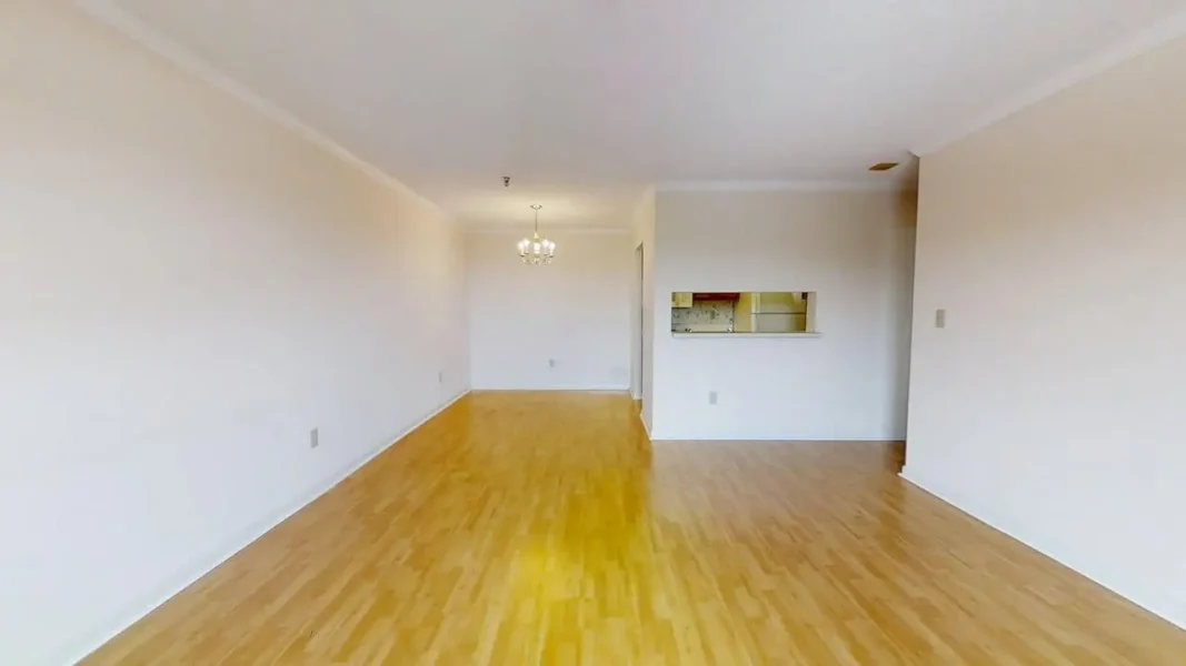 Empty living room with warm laminate flooring, white walls, and a chandelier overhead; a kitchen pass-through window is visible on the right side.