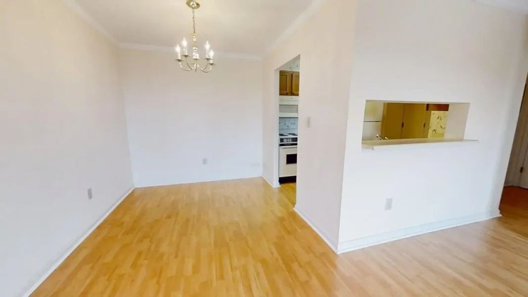 Empty dining area with a chandelier, light hardwood floors, and a kitchen pass-through window in a bright, white-walled room