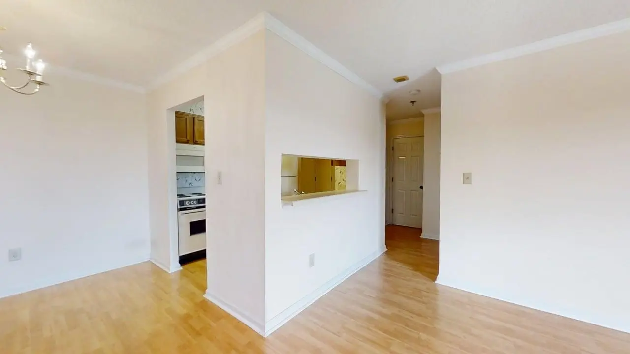 Bright empty living area with a half-wall pass-through to the kitchen, doorway to a hallway, and light wood floors.