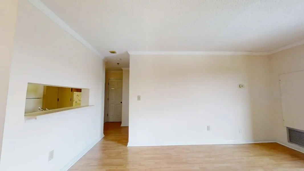 Empty living area with light walls, wood flooring, and a cutout pass-through to a kitchen on the left; doorway at the far end.