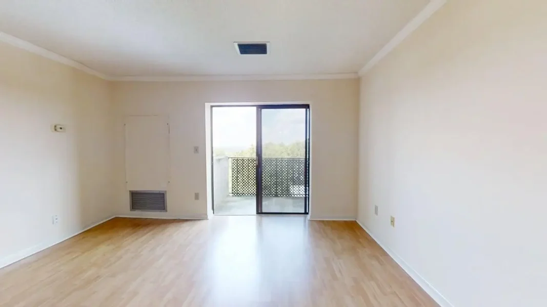 Empty living room with sliding glass door to a balcony, light wood flooring, and beige walls; minimal and bright.