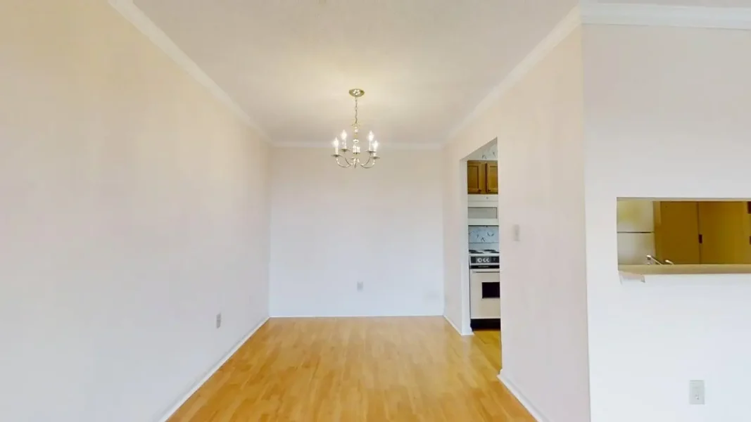 Bright, empty dining area with a hanging chandelier and light wood flooring; pass-through to a kitchen with a stove and cabinets visible through a doorway.