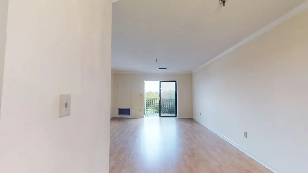 Empty living room with light wood flooring, off-white walls, and a sliding glass door to a balcony.