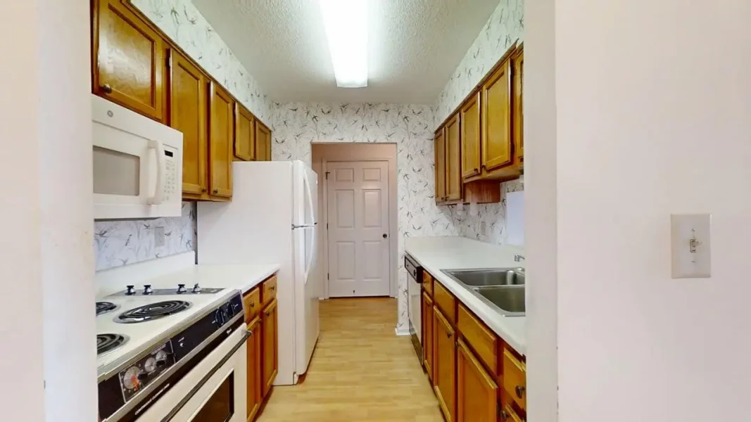 Narrow galley kitchen with white appliances, oak cabinets, and a double sink along the right side; stove and microwave on the left.