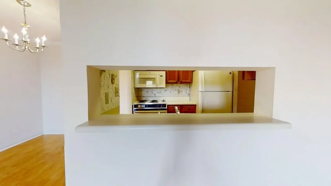 View into a small kitchen through a rectangular pass-through in a white wall; stove, microwave, fridge and wooden cabinets are visible.