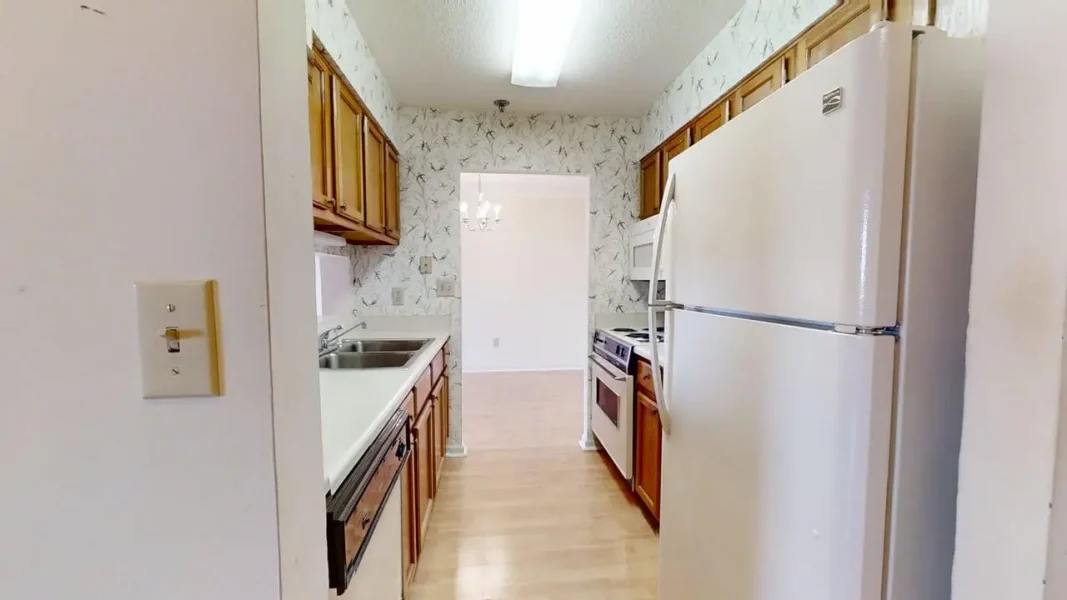 Narrow galley kitchen with white appliances, wooden cabinets, and light wood flooring leading to a dining area.