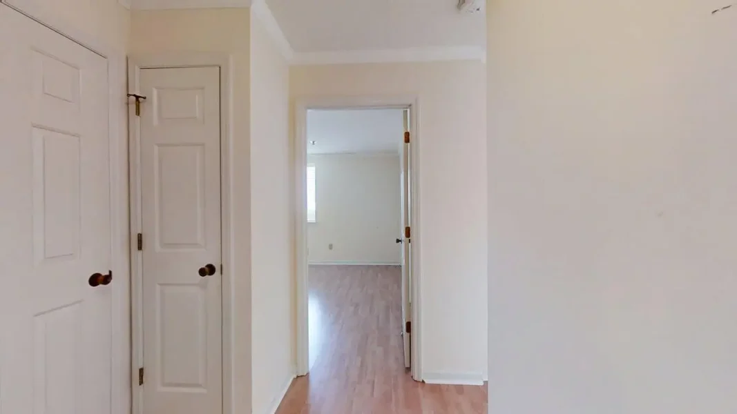 Bright hallway with white doors and light wood flooring, open doorway at the end leading to a sunlit room.