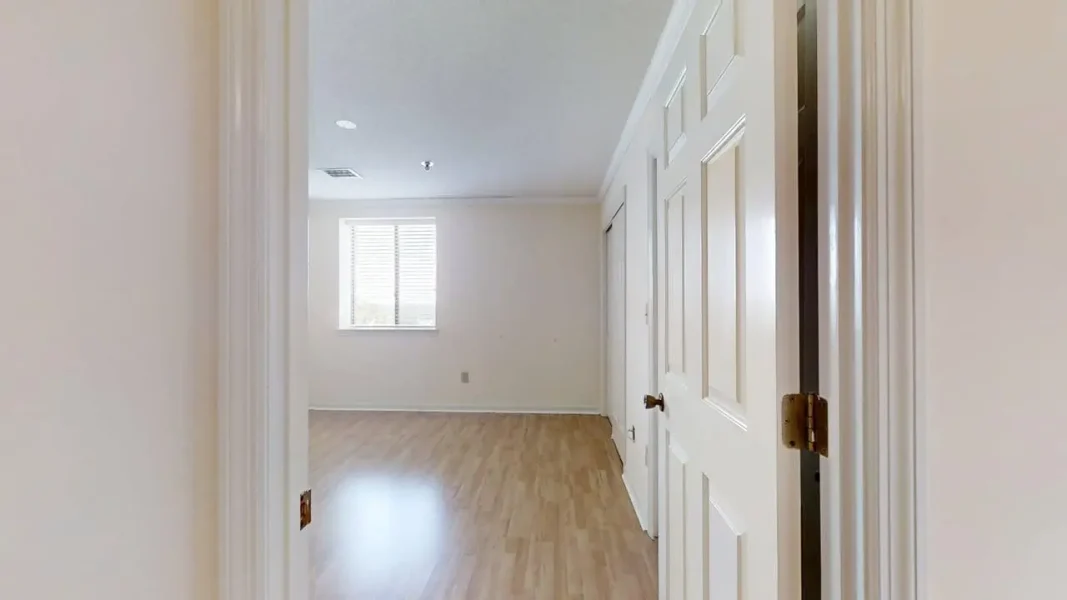 View from a doorway into an empty room with light wood flooring, white walls, a single window with blinds, and several white doors along the right wall.