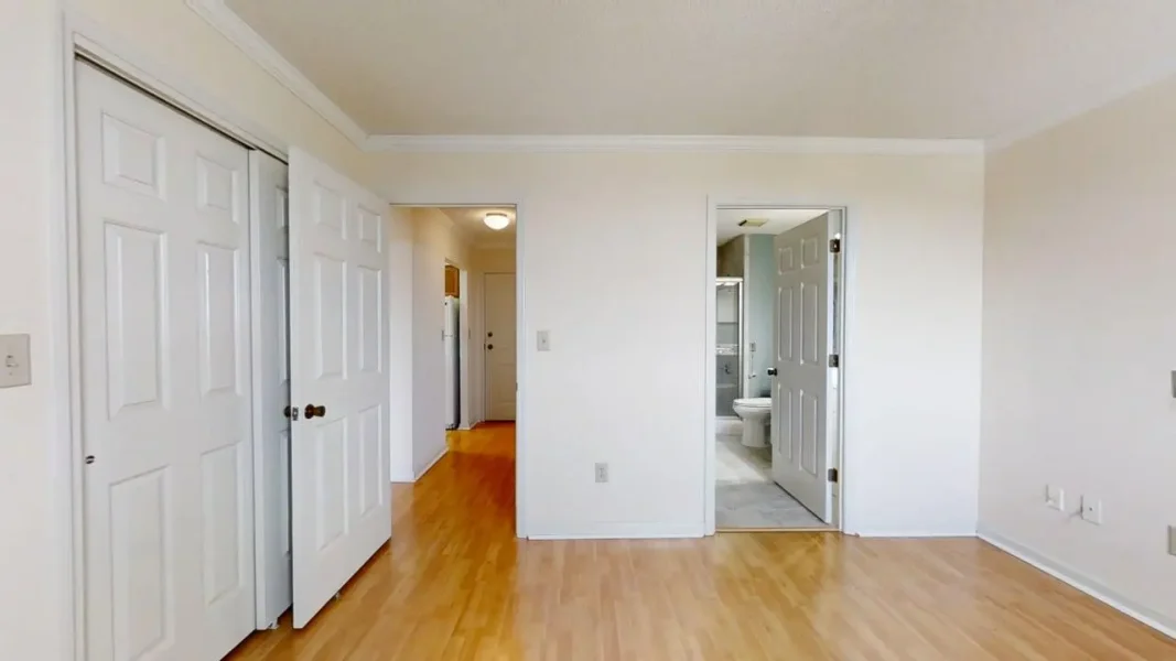Bedroom with white walls, light wood flooring, and two closet doors on the left; an open doorway leads to a hallway and another open door reveals a bathroom with a visible toilet.
