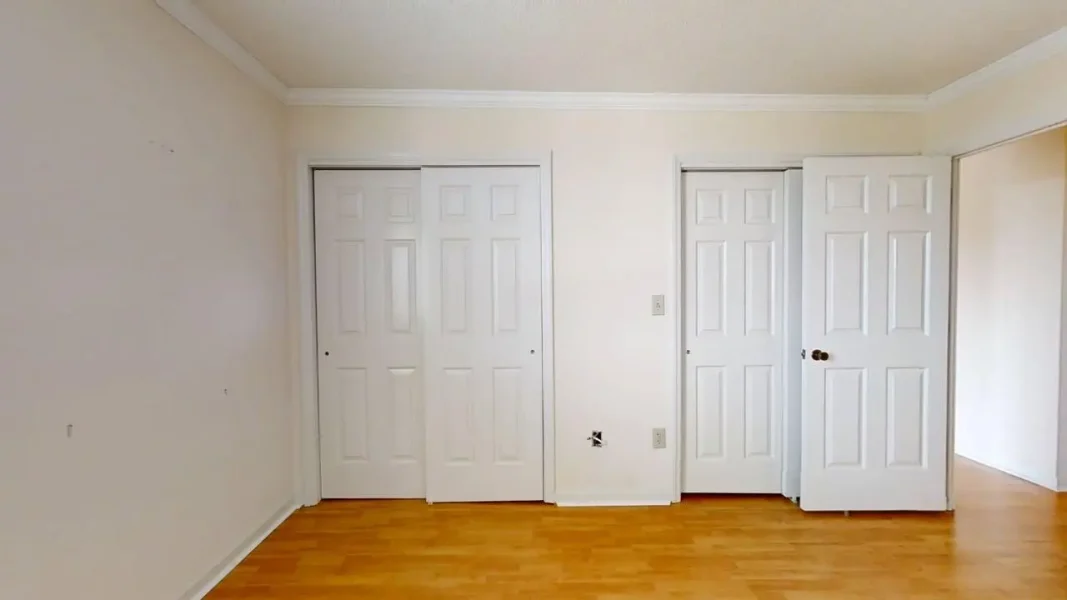 Empty room with light wood flooring and three white doors along the back wall (two closet doors and one entry door). The space is bare, with a switch and outlet visible on the wall.